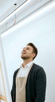 Young man with light brown hair and beard, Caucasian ethnicity, wearing beige apron over white shirt and dark cardigan, smiling while looking upward in bright modern interior space.