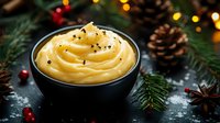 Creamy mashed potatoes topped with black pepper in dark bowl, surrounded by pine cones, fir branches and holiday lights on rustic background.