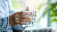 Senior man holding glass of water with pill, close up view of elderly hands with medication and clear drinking glass against blurred background. Healthcare concept.
