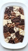 Assorted chocolate pieces with nuts and toppings arranged on white plate, featuring dark, milk, and white varieties for confectionery, dessert, and food styling projects.