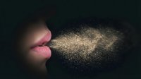 Female lips blowing golden glitter particles against dark background creating magical sparkle effect for cosmetics and beauty concepts.