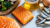 Overhead view of fresh salmon fillet, sardines, broccoli, eggs, and vitamin oil bottle arranged on white surface for healthy nutrition and wellness content.
