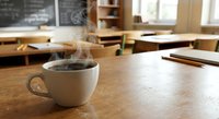 Steaming hot coffee cup on wooden desk in bright classroom with chalkboard, books and pencils creating cozy academic learning environment.