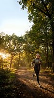 Young woman jogging through sunlit forest path during golden hour morning workout in natural park setting for healthy lifestyle and fitness.