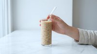 Hand holding glass of quinoa with spoon on white marble table, healthy grain food for nutrition and diet planning, cooking ingredient for wellness recipes.