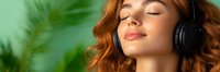 Young caucasian woman with red hair and closed eyes enjoying music in wireless headphones against green blurred nature background, relaxation and mindfulness concept.
