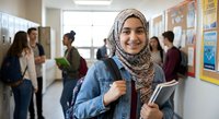 Young Muslim female student wearing hijab smiling in school hallway holding books and backpack with diverse classmates in background.