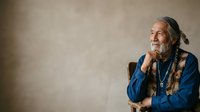 Mature man with long gray hair and beard, wearing blue shirt and necklace, sitting in contemplative pose with hand to chin against neutral background.
