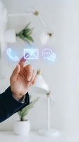 Hand touching digital communication icons with phone, email and chat symbols floating above palm in modern office environment.