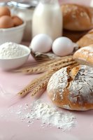 Fresh baked bread with flour, wheat ears, eggs, milk and baking ingredients on pink background, rustic food composition for bakery or cooking concept.