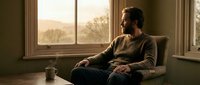 Thoughtful bearded man relaxing in armchair by window during golden hour sunset, enjoying peaceful moment with coffee cup in cozy home interior.