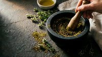 Hand grinding fresh herbs and spices in traditional stone mortar and pestle for cooking preparation on rustic kitchen counter with olive oil.