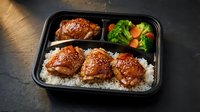 Glazed teriyaki chicken thighs over steamed rice with fresh broccoli and carrots in black takeout container for meal prep and healthy dining options.