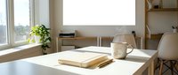 Minimalist workspace with white desk, open notebook, ceramic mug, and potted plant by window with natural light, suitable for office and productivity concepts.