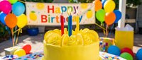 Bright yellow birthday cake with colorful candles at an outdoor party, surrounded by balloons and a festive Happy Birthday banner.