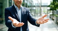 Professional businessman in navy suit with open welcoming hands gesture in modern office building lobby for business presentations and corporate communication.
