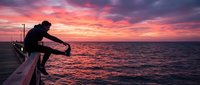 Young man stretching on a wooden pier at dramatic pink and red sunset over calm ocean, silhouette against vibrant sky.