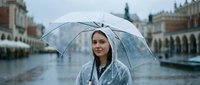 Young woman with fair complexion and long dark hair holding transparent umbrella in rainy weather at historic European square with classical architecture, wearing casual jacket, suitable for travel, weather, lifestyle and tourism content.