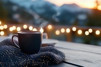 Steaming coffee cup on knitted scarf against blurred mountain sunset and bokeh lights background, creating cozy winter atmosphere on wooden deck.