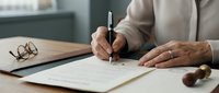 Professional businessman signing important legal contract document at modern office desk with pen and glasses nearby for business agreement.