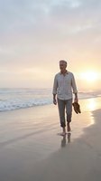 Mature man with gray hair and light complexion walking barefoot on sandy beach at sunset, holding shoes in hand, wearing casual light blue shirt and khaki pants, golden hour lighting.