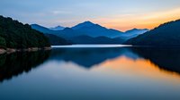 Tranquil mountain lake at sunset with perfect mirror reflection of silhouetted peaks and golden sky, creating serene landscape with smooth water surface.