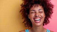 Young African American woman with natural curly hair laughing joyfully against colorful yellow and pink background, expressing genuine happiness and positive emotions.