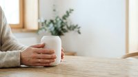Person holding warm white mug on wooden table in bright minimalist interior with potted plant and window, suitable for lifestyle and wellness content.