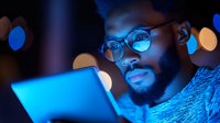 Young African American man in glasses illuminated by blue light from digital tablet screen at night, with bokeh lights creating atmospheric background effect.