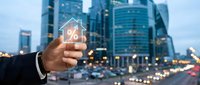 Businessman hand holding glowing house icon with percentage symbol over blurred city skyline at dusk. Real estate investment and mortgage rates concept.