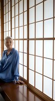 Senior woman with white hair wearing blue shirt leaning against wooden grid window with natural light, suitable for lifestyle, wellness, and aging gracefully concepts.