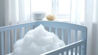 Light blue wooden baby crib with white fluffy cloud-shaped pillow in bright nursery room with natural window light for infant sleep comfort.