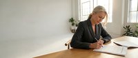 Senior woman with gray hair, Caucasian ethnicity, wearing dark business blazer, signing documents at wooden desk in bright office with windows and plants.