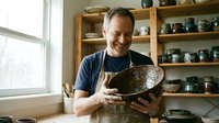 Mature male artisan with dark hair and facial hair, wearing navy apron, examining handcrafted ceramic bowl in pottery studio with wooden shelving and ceramic vessels. Suitable for craft business, artisan workshops, and creative professional content.