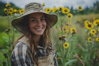 Young female farmer in sun hat and overalls smiling in sunflower field during summer harvest, authentic countryside lifestyle perfect for agricultural and farming campaigns.