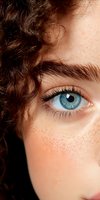 Close-up view of bright blue eye with natural freckles and curly brown hair, showing detailed iris pattern and long eyelashes against soft skin texture.