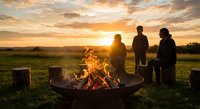 Friends gathering around outdoor fire pit during golden hour sunset in countryside field with warm flames and dramatic sky.