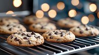 Freshly baked chocolate chip cookies cooling on black wire rack with warm bokeh lights in background, selective focus on golden brown treats.