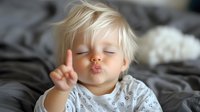 Playful toddler with blonde hair lying on bed making peace sign and kiss face gesture, wearing patterned white shirt. Sweet candid childhood moment.