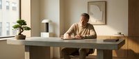Mature man with light skin tone, gray hair, wearing a beige button-up shirt, seated at a minimalist desk in a modern office workspace with natural lighting, potted plant, and contemporary decor for business and professional concepts.