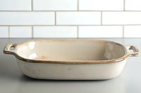 Vintage white ceramic baking dish with brown rim and handles on marble counter against subway tile wall, rustic farmhouse kitchen cookware.