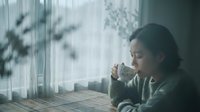 Young Asian woman in cozy sweater drinking hot beverage by window with natural light and shadow patterns creating peaceful morning atmosphere at home.