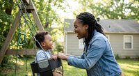 African American father pushing young son on swing in backyard playground, family bonding time outdoors with residential home background.