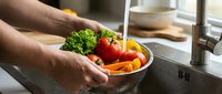 Fresh vegetables being washed in kitchen sink with running water. Healthy cooking preparation with broccoli, tomatoes and carrots for nutritious meal.