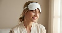 Young Caucasian woman wearing white sleep mask and beige robe relaxing with closed eyes in bright bedroom. Self-care wellness routine concept.