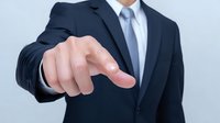 Businessman in navy suit pointing finger forward against light background, demonstrating leadership and decision-making in corporate environment.