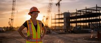 Young Hispanic male construction worker in safety vest and hard hat standing confidently at building site during golden hour sunset with cranes in background.