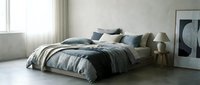 Modern bedroom with unmade bed featuring striped gray and white bedding, decorative pillows, wooden nightstands, and minimalist decor in bright natural light.