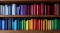 Colorful books arranged by color spectrum on wooden shelves creating rainbow gradient effect, spines facing outward for decorative library display.
