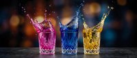 Colorful liquid splashing from crystal glasses with pink blue yellow drinks creating dynamic motion against blurred bokeh background for beverage advertising.